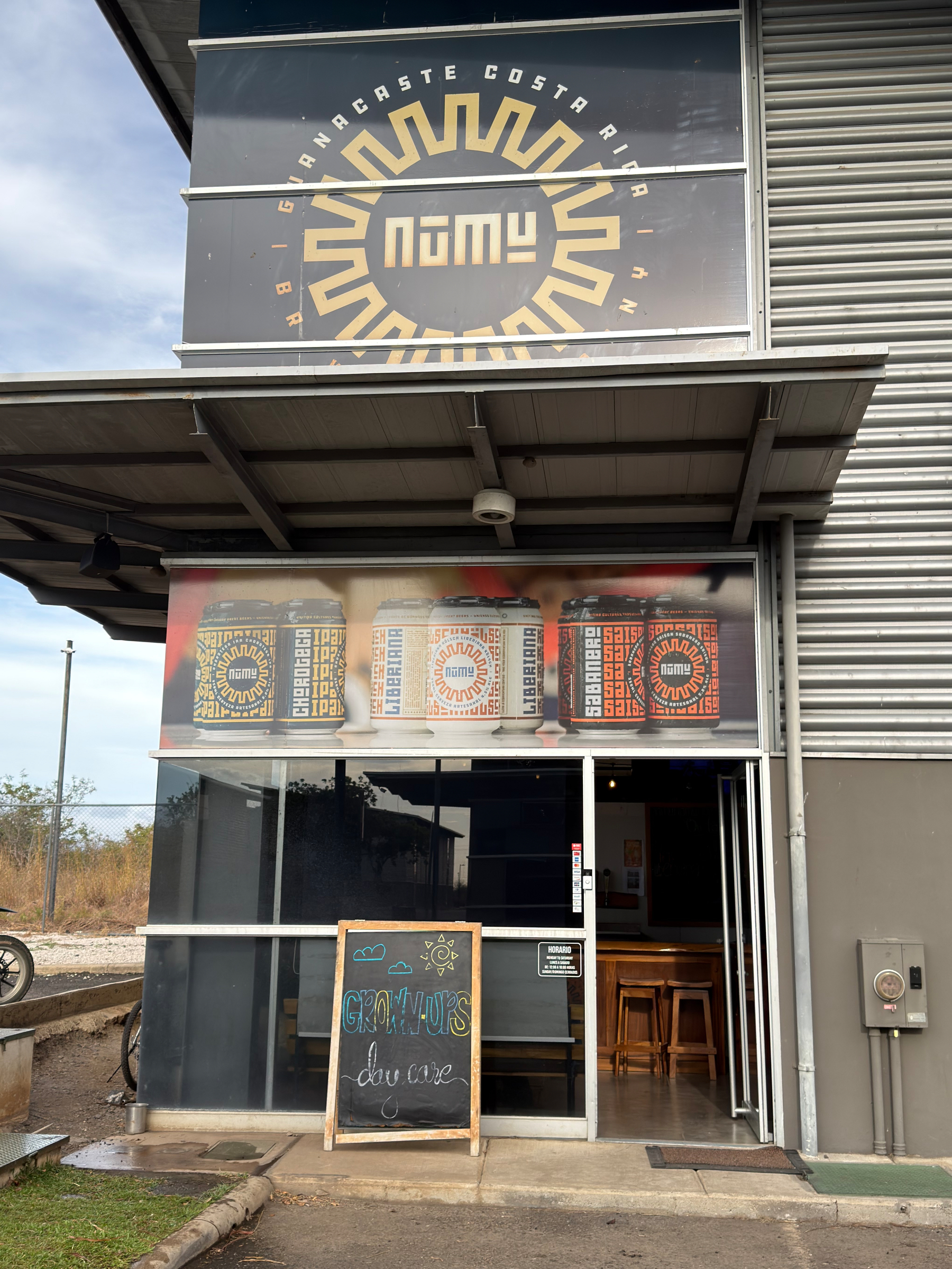 An industrial-style building has a sign displaying the business's logo, with a chalkboard outside and an open entrance leading to a seating area.